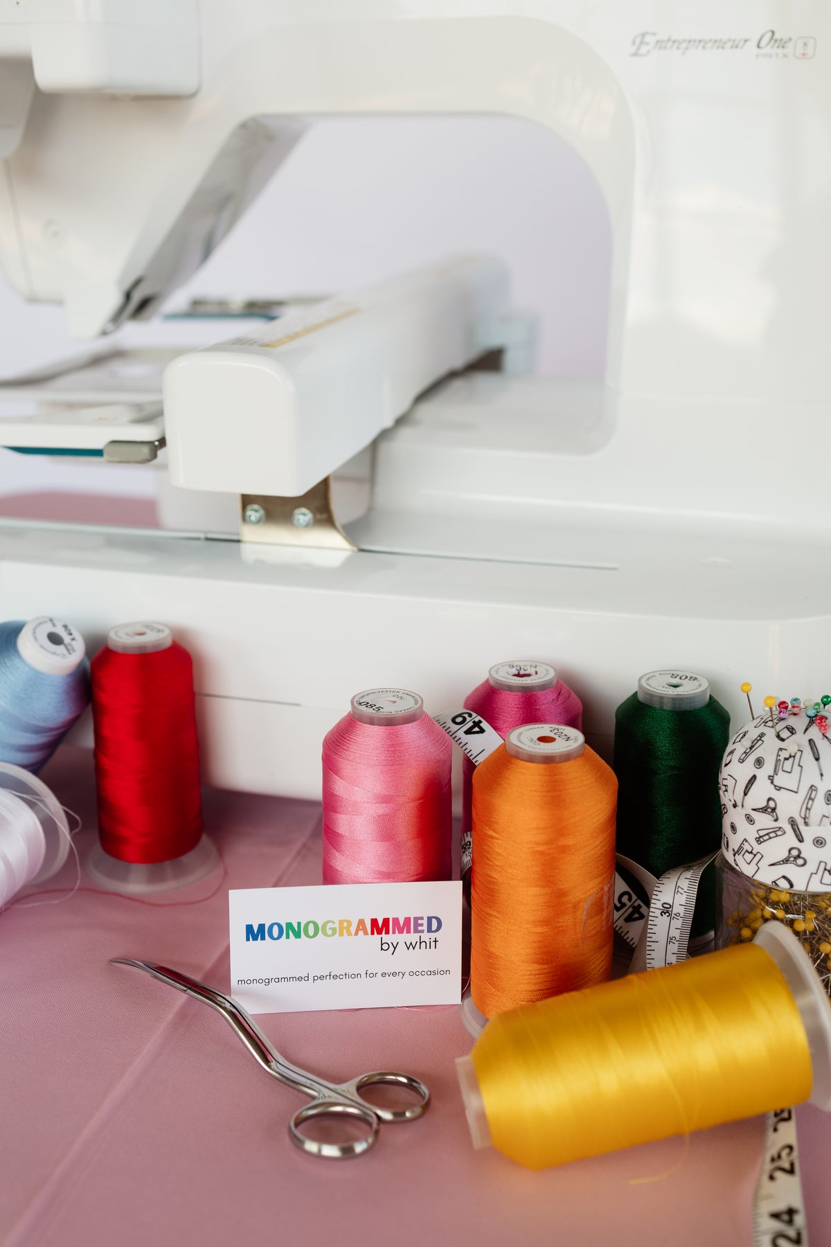 Spools of embroidery thread in pink, orange, green and blue beside an embroidery machine at a live event.