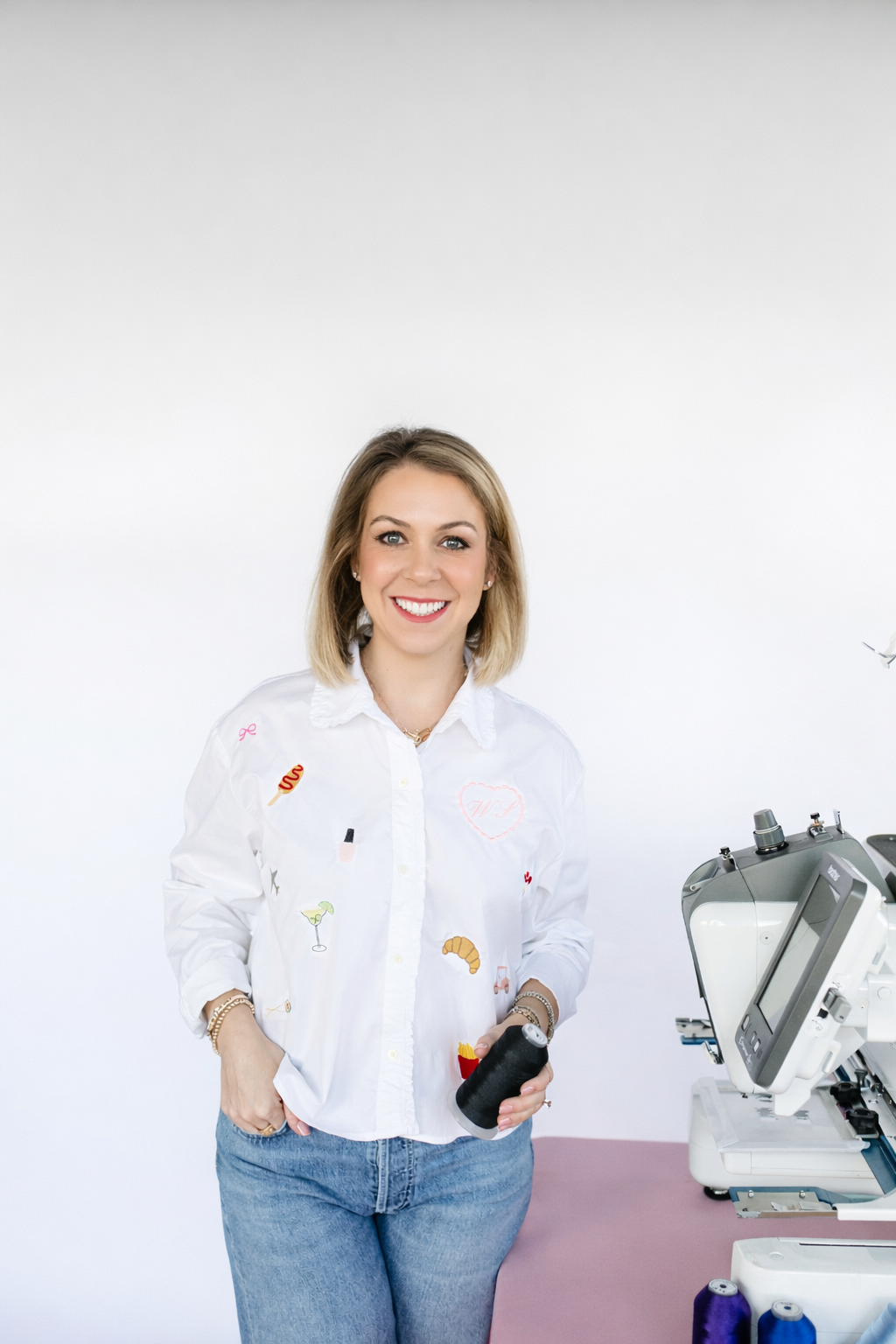 Whitney standing beside her embroidery machine, holding a spool of thread and wearing a white shirt covered in small embroidered motifs.