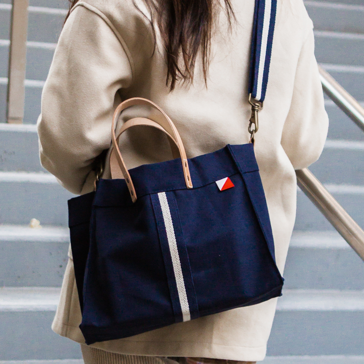 Woman carrying the mini navy canvas box tote with leather handles and a navy and white striped crossbody strap