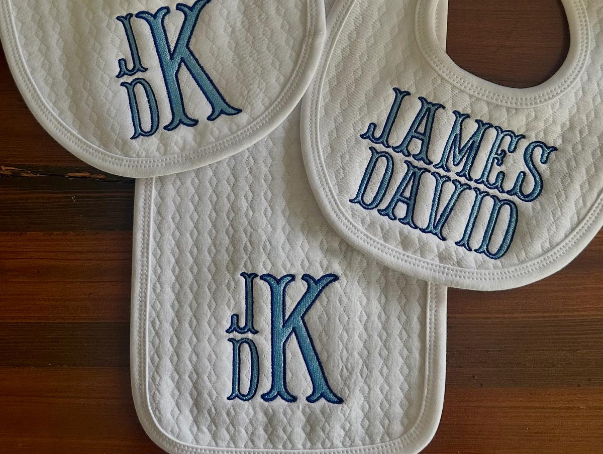 Three white quilted baby bibs and a burp cloth, monogrammed in blue thread with a stacked JdK monogram and the name James David.