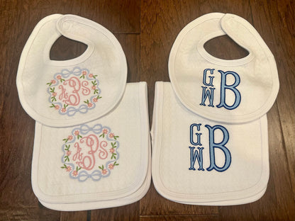Two white baby bib and burp cloth sets, one monogrammed with a pink floral wreath monogram and one with a blue GMB stacked monogram