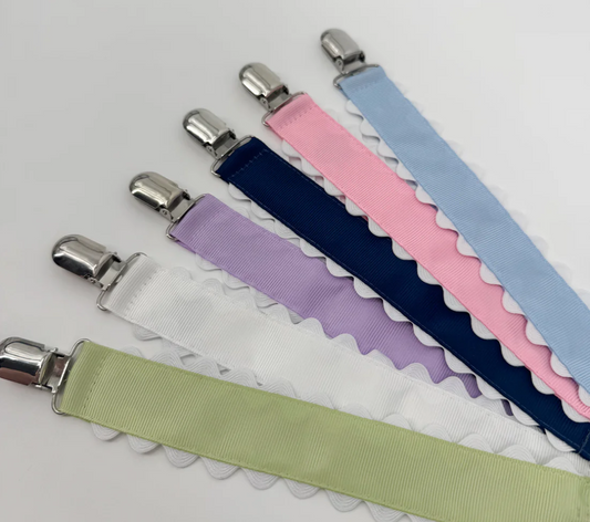 Six ribbon pacifier clips with white ric rac trim, fanned out in light blue, pink, navy, white, purple, and green