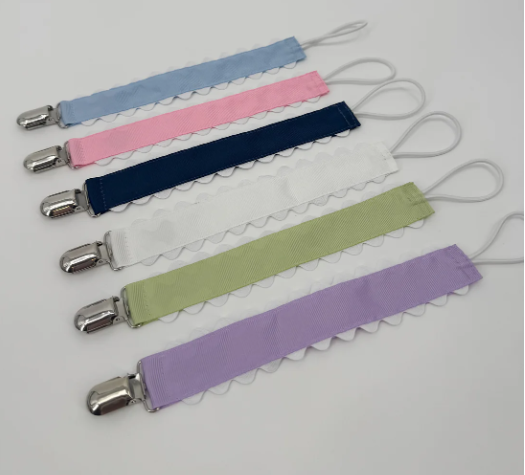 Six ribbon pacifier clips with white ric rac trim and metal clips laid out in a row in light blue, pink, navy, white, green, and purple