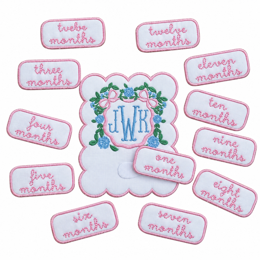 Embroidered monthly milestone card with a blue JWK monogram in a hydrangea wreath with a pink bow, surrounded by pink embroidered month patches